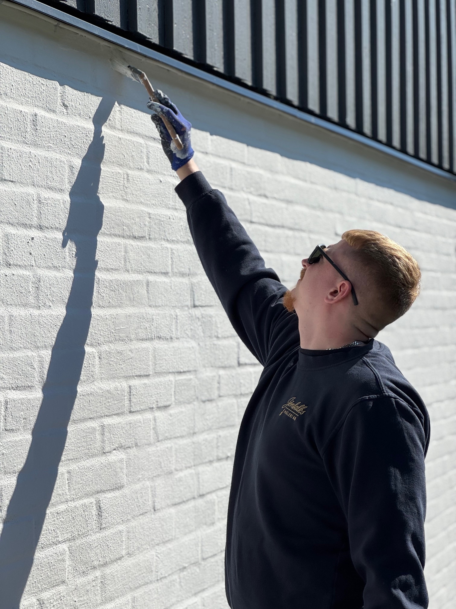 En professionell målare i vit arbetsdräkt målar en husfasad från en byggställning. Med pensel och noggrannhet arbetar han med utvändig målning runt en ventilation. Perfekt exempel på fasadmålning och renovering av hus.